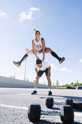 Woman jumping over man on street against sky in city during sunny day - JCMF01127