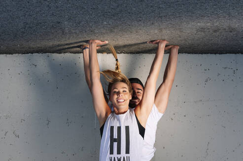 Smiling couple practicing handstands on road against wall in city - JCMF01131