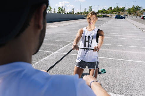 Couple exercising with strap while standing on road in city during sunny day - JCMF01132