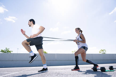 Couple exercising with strap on road against sky during sunny day - JCMF01136