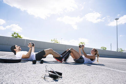 Couple holding dumbbells while lying on road against sky during sunny day - JCMF01140