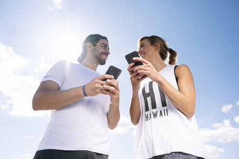 Smiling couple holding smart phones looking at each other while standing against sky during sunny day - JCMF01142