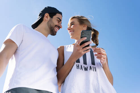 Smiling woman holding smart phone looking at boyfriend against clear blue sky - JCMF01143