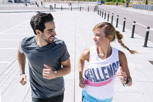 Smiling couple looking at each other while running on sidewalk in city - JCMF01144