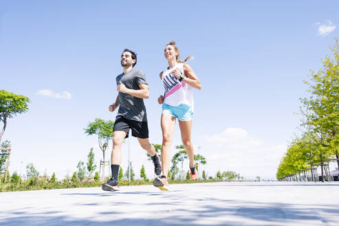 Mid adult couple running on road against blue sky during sunny day - JCMF01149