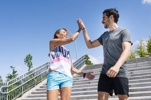 Cheerful couple giving high-five while standing on steps against blue sky in city - JCMF01152