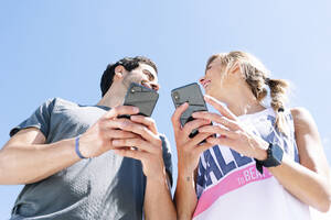 Lächelndes Paar, das sich gegenseitig ansieht, während es ein Smartphone gegen den klaren blauen Himmel benutzt - JCMF01156