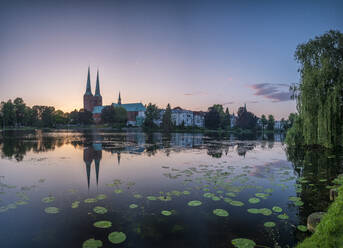 Germany, Schleswig-Holstein, Lubeck, Water lilies growing on bank of Trave at dusk with old town buildings in background - HAMF00695