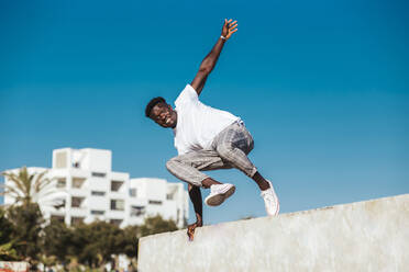 Afrikanischer Mann springt über Stützmauer gegen klaren blauen Himmel an einem sonnigen Tag - LJF01692