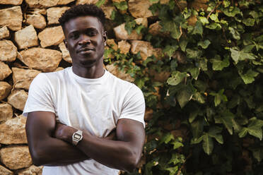 Young man with arms crossed standing against stone wall - LJF01697