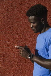 Nahaufnahme eines jungen Mannes, der sein Smartphone an einer roten Wand in einer Stadt an einem sonnigen Tag benutzt - LJF01701
