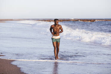 Shirtless African young man running at shore against clear sky - LJF01707