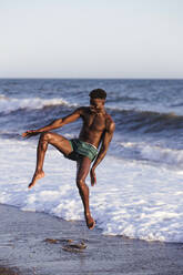 Shirtless African young man jumping at shore against clear sky - LJF01708