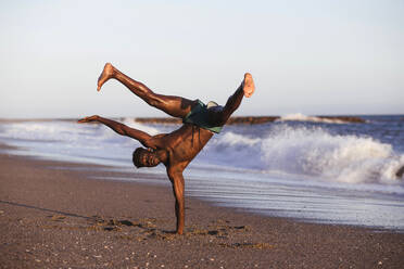 Shirtless African man doing handstand at beach against clear sky - LJF01711