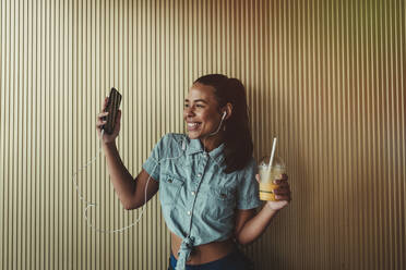 Cheerful woman holding smart phone and drink while enjoying music through headphones against wall - DSIF00065