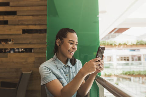 Fröhliche junge Frau, die ihr Smartphone benutzt, während sie an einem Geländer in einem Einkaufszentrum sitzt, lizenzfreies Stockfoto