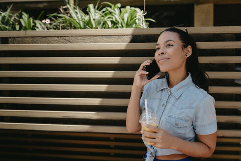 Smiling young woman holding juice talking over smart phone against wall in city - DSIF00070