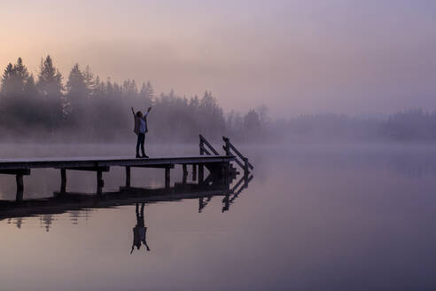 Woman standing with raised arms on lakeshore jetty at foggy dawn - LBF03170