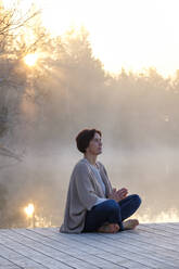 Adult woman meditating on lakeshore jetty at foggy sunrise - LBF03171