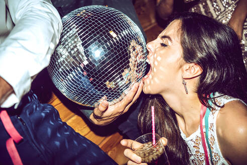 Young woman with eyes closed holding drink and biting disco ball in glamorous party - EHF00666