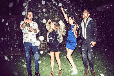 Young man opening champagne while friends dancing outdoors in party at night - EHF00693