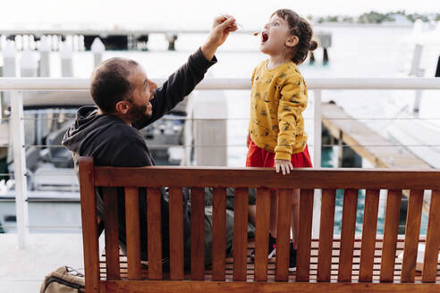 Vater füttert Tochter auf einer Bank am Meer in der Stadt mit Limettenkuchen - GEMF03944