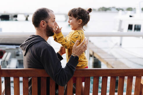 Vater spielt mit fröhlicher Tochter auf einer Bank am Meer in der Stadt - GEMF03945