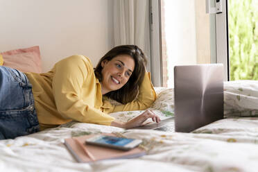 Smiling woman using laptop while lying in bedroom - AFVF06843