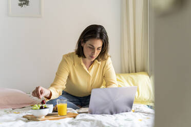 Frau isst Frühstück und benutzt Laptop im Schlafzimmer - AFVF06849