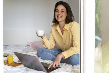 Lächelnde Frau, die einen Kaffee trinkt und einen Laptop im Schlafzimmer benutzt - AFVF06851
