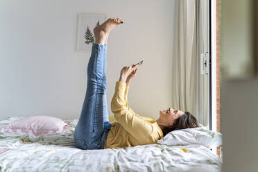 Smiling woman using smart phone while lying with feet up in bedroom - AFVF06855