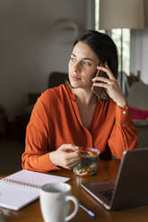 Businesswoman talking on mobile phone while eating salad at home - AFVF06882