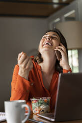 Happy businesswoman talking on mobile phone while eating salad at home - AFVF06883