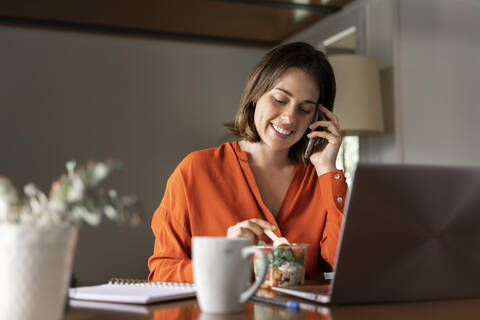 Smiling businesswoman talking on smart phone while eating salad at home stock photo