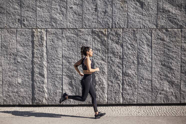 Beautiful and brunette woman running on a sunny morning with a granite wall in the background - ADSF07120