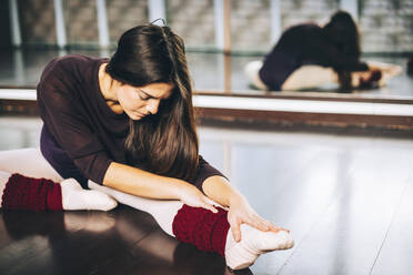 Mädchen übt Ballett und spannt Muskeln an, während sie auf dem Boden des Studios sitzt. - ADSF07181