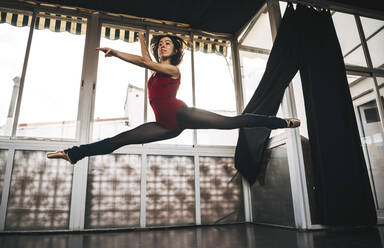 Young slim ballerina jumping above ground in studio flexing legs. - ADSF07191