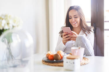 Attractive young happy woman using mobile phone and having breakfast at table near window at home - ADSF07198