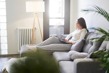 Seitenansicht einer attraktiven jungen glücklichen Frau, die einen Laptop benutzt und sich zu Hause auf dem Sofa ausruht - ADSF07200