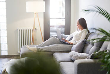 Side view of attractive young happy woman using laptop and resting on sofa at home - ADSF07200