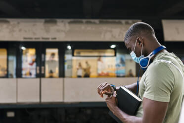 Young man wearing mask checking time while standing at subway station - EGAF00602