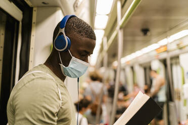 Nahaufnahme eines jungen Mannes mit Maske, der im Zug ein Buch liest und über Kopfhörer Musik hört - EGAF00603