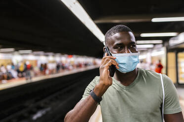 Young man wearing mask talking over smart phone while standing at subway station - EGAF00608