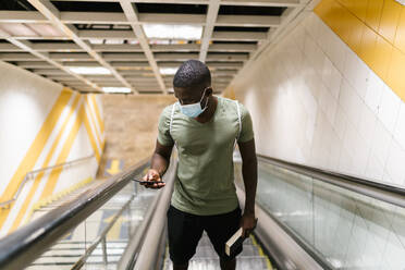 Junger Mann mit Maske benutzt sein Smartphone, während er auf einer Rolltreppe in einer U-Bahn-Station steht - EGAF00610