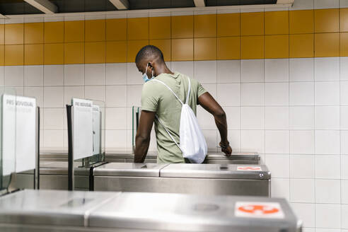 Junger Mann mit Maske und Rucksack geht durch ein Drehkreuz in einer U-Bahn-Station - EGAF00611