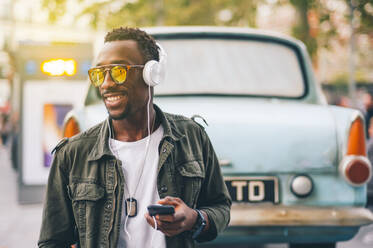 Lächelnder junger Mann mit Sonnenbrille hört Musik über Kopfhörer gegen Oldtimer in der Stadt - OCMF01578