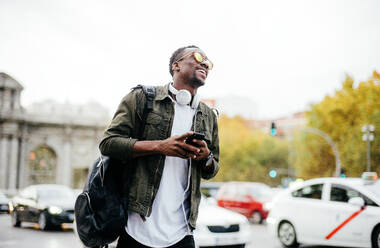Glücklicher Mann mit Sonnenbrille, der sein Smartphone in der Hand hält und gegen den Himmel in der Stadt steht - OCMF01580