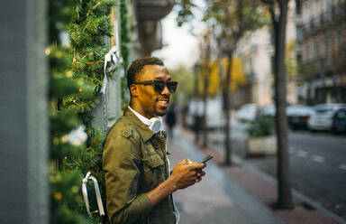 Junger Mann mit Sonnenbrille, der sein Smartphone benutzt, während er auf dem Bürgersteig in der Stadt steht - OCMF01582
