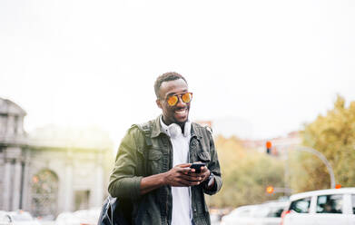 Smiling man wearing sunglasses using smart phone while standing against clear sky in city - OCMF01583