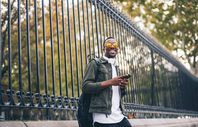 Smiling young man wearing sunglasses using mobile phone while standing against fence in city - OCMF01587